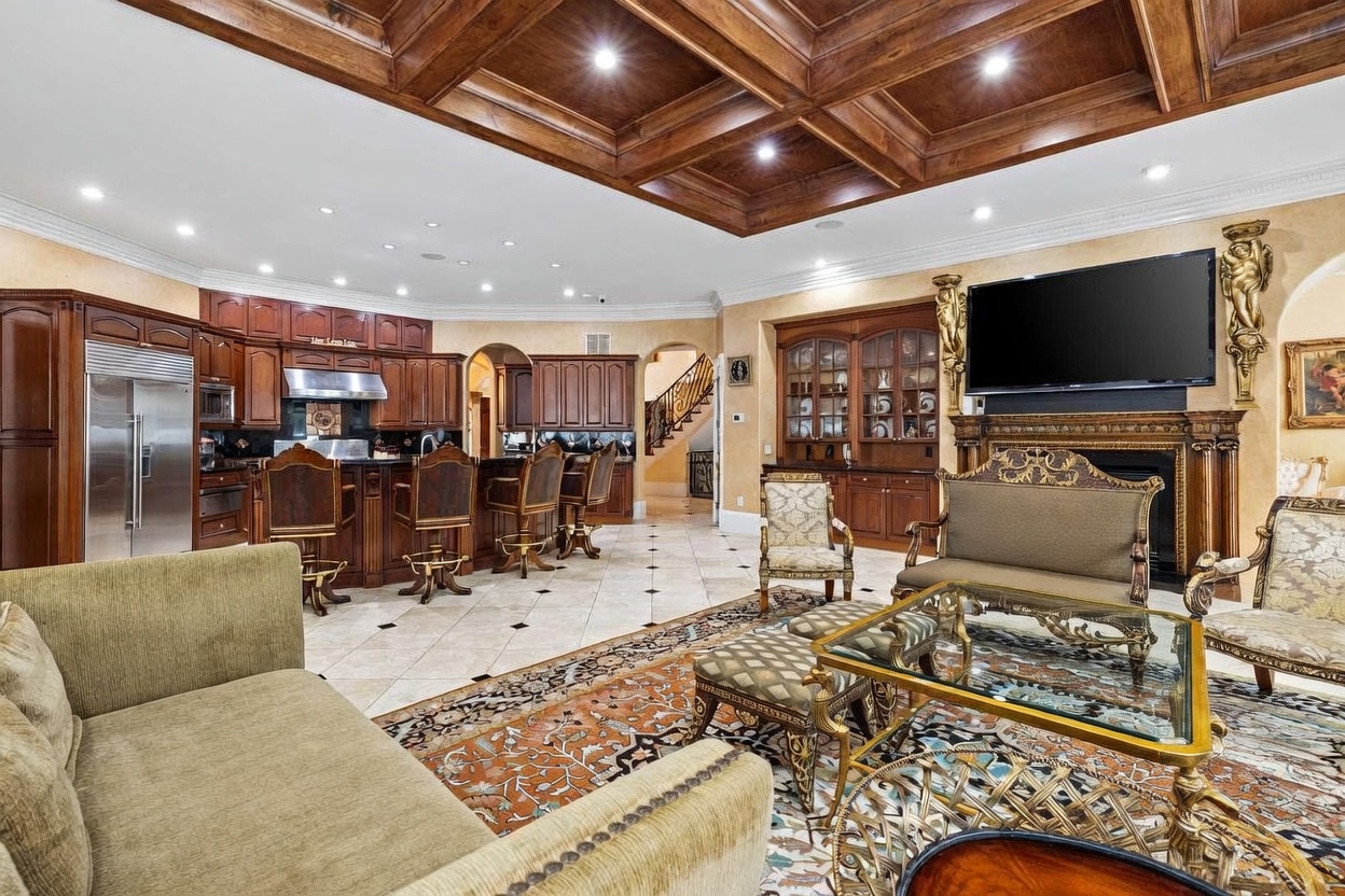 This expansive open-concept living area features a coffered wood ceiling and ornate, traditional decor. The space includes a formal seating area with a television mounted above a fireplace, a kitchen with a breakfast bar, and a view leading toward a staircase in the background. The room is finished with patterned tile flooring and a large area rug, creating a cohesive and luxurious atmosphere.
