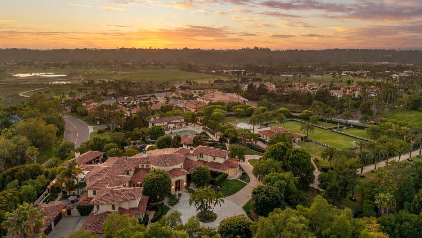 This aerial view showcases a sprawling estate with a red-tiled roof, surrounded by lush greenery and mature trees. The property features a circular driveway, manicured lawns, and what appears to be a tennis court and pool. The overall impression is one of luxury and privacy, set against a scenic backdrop with a beautiful sunset.