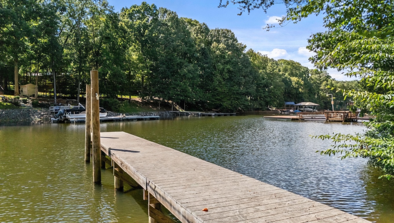 This serene waterfront view features a long wooden dock extending into a calm, tree-lined lake. The lush, dense forest surrounding the water creates a private and tranquil atmosphere, with additional docks visible in the distance. The perspective is from the end of the dock, looking out across the water toward the wooded shoreline.