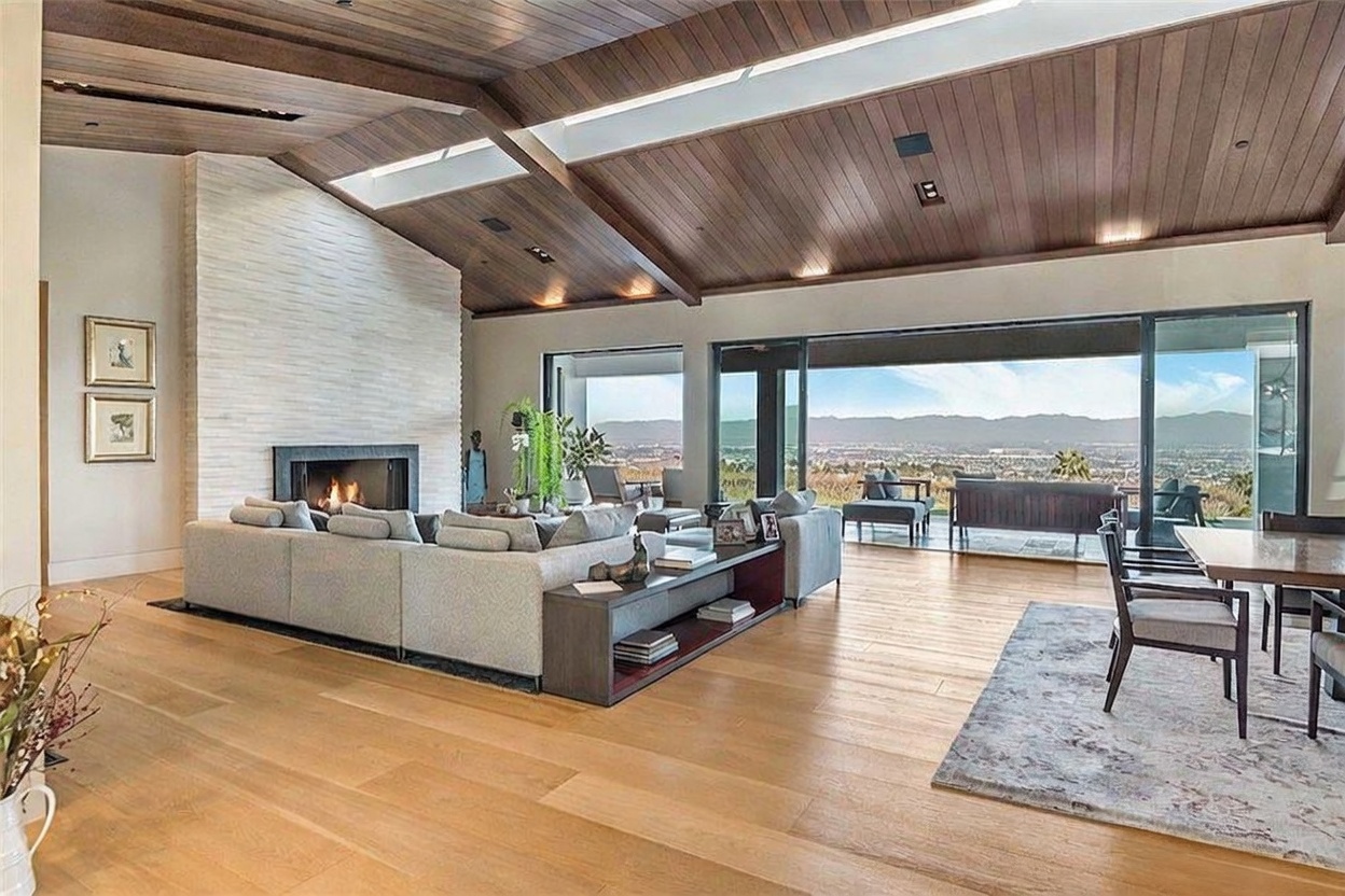 This expansive open-concept living area features a vaulted wood-paneled ceiling with integrated skylights and a prominent stone-clad chimney breast. A large sectional sofa faces the fireplace, while a wall of sliding glass doors provides an unobstructed panoramic view of the distant mountains and valley. The space is finished with light wood flooring and a neutral color palette, creating a bright and airy atmosphere.