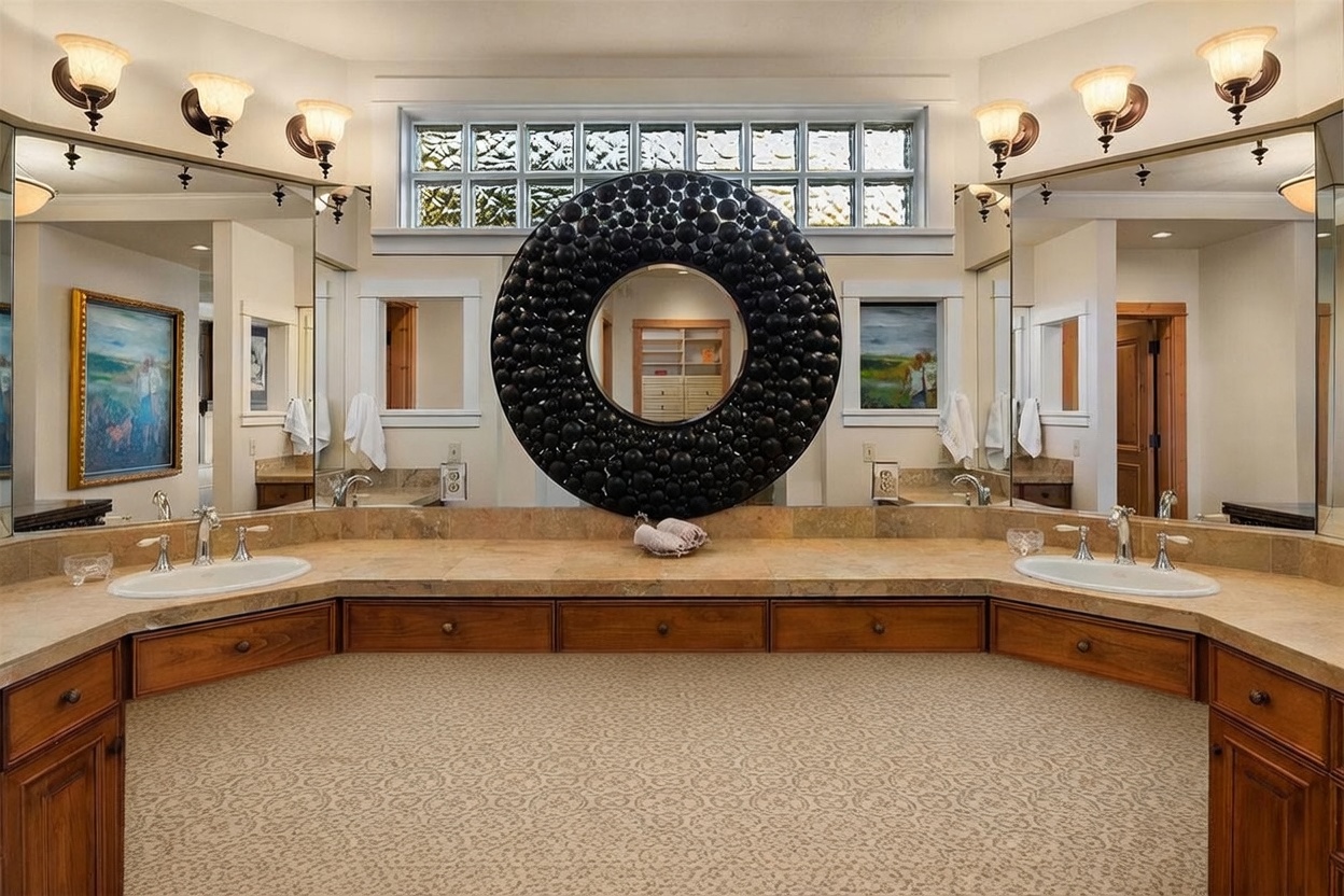 This primary bathroom features a dual-sink vanity with warm-toned wood cabinetry and a neutral stone countertop. A striking, circular black textured mirror serves as the focal point between two large wall mirrors, all positioned beneath a row of glass block windows. The space is illuminated by multiple wall-mounted light fixtures, creating a symmetrical and traditional aesthetic.