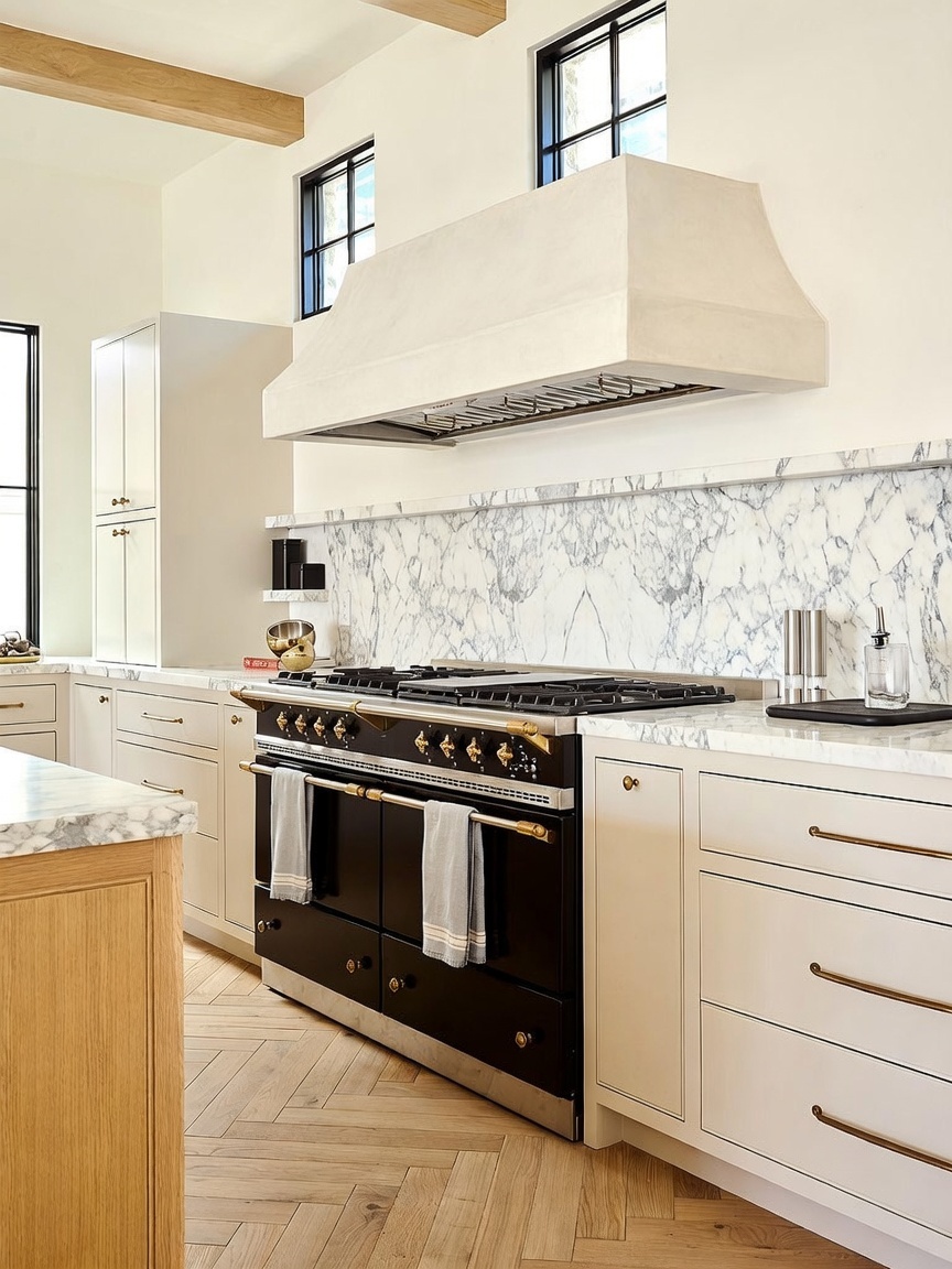 This elegant kitchen features a prominent black range with gold-toned hardware set beneath a custom-designed, light-colored range hood. The space is accented by a striking stone backsplash with dramatic veining, light-toned cabinetry, and herringbone-patterned wood flooring. The perspective captures the cooking area, highlighting the blend of classic and modern design elements.