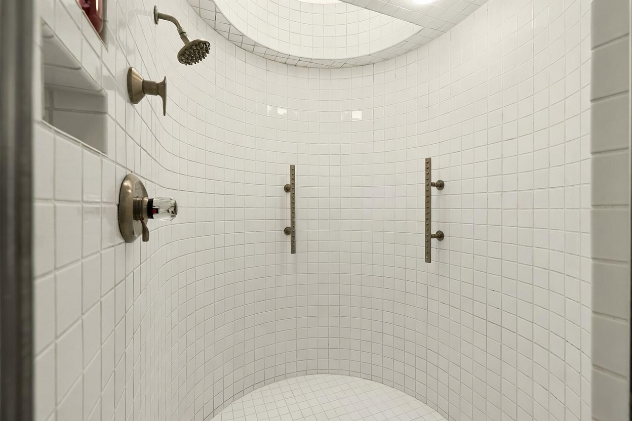 This curved shower enclosure features white square tiles covering the walls and floor, creating a clean and minimalist aesthetic. The space is equipped with a wall-mounted shower head, a separate handheld sprayer, and two vertical grab bars for safety. A recessed niche is built into the wall for storage, and the curved design adds a unique architectural element to the bathroom.