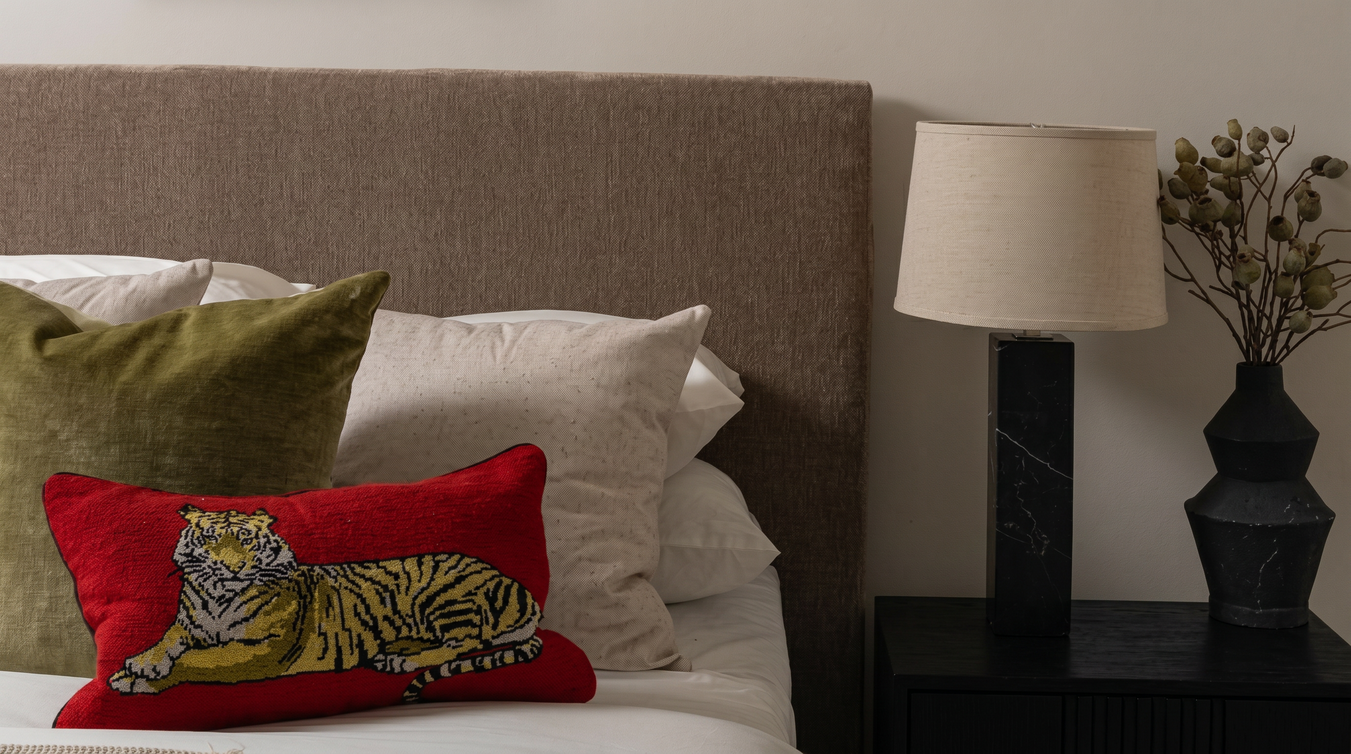 This cozy bedroom scene features a textured brown upholstered headboard accented by a layered display of pillows, including a distinctive red tiger-patterned throw pillow. To the side, a sleek black marble-base table lamp stands atop a dark wooden nightstand, paired with a modern matte black vase containing dried foliage. The composition creates an inviting, sophisticated, and earthy atmosphere.