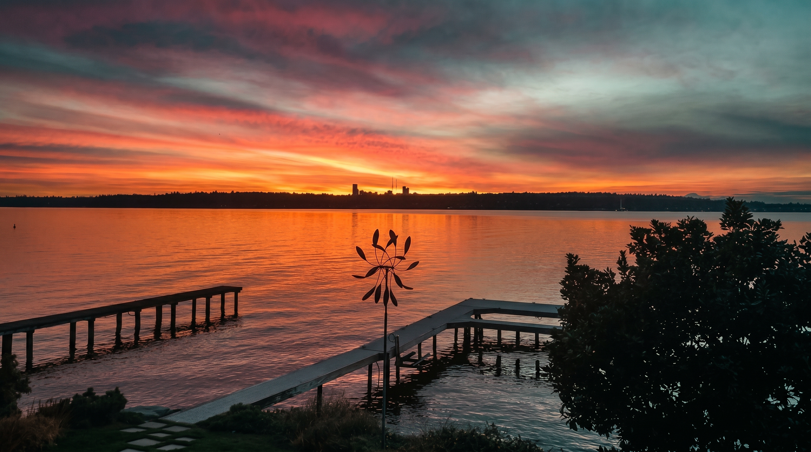 This scenic waterfront view captures a vibrant sunset over a calm body of water, with a distant city skyline silhouetted against the fiery orange and purple sky. In the foreground, a private wooden dock extends into the water, accompanied by a decorative metal wind spinner set within a landscaped yard. The perspective is elevated, looking out from the property toward the horizon, creating a serene and picturesque atmosphere.