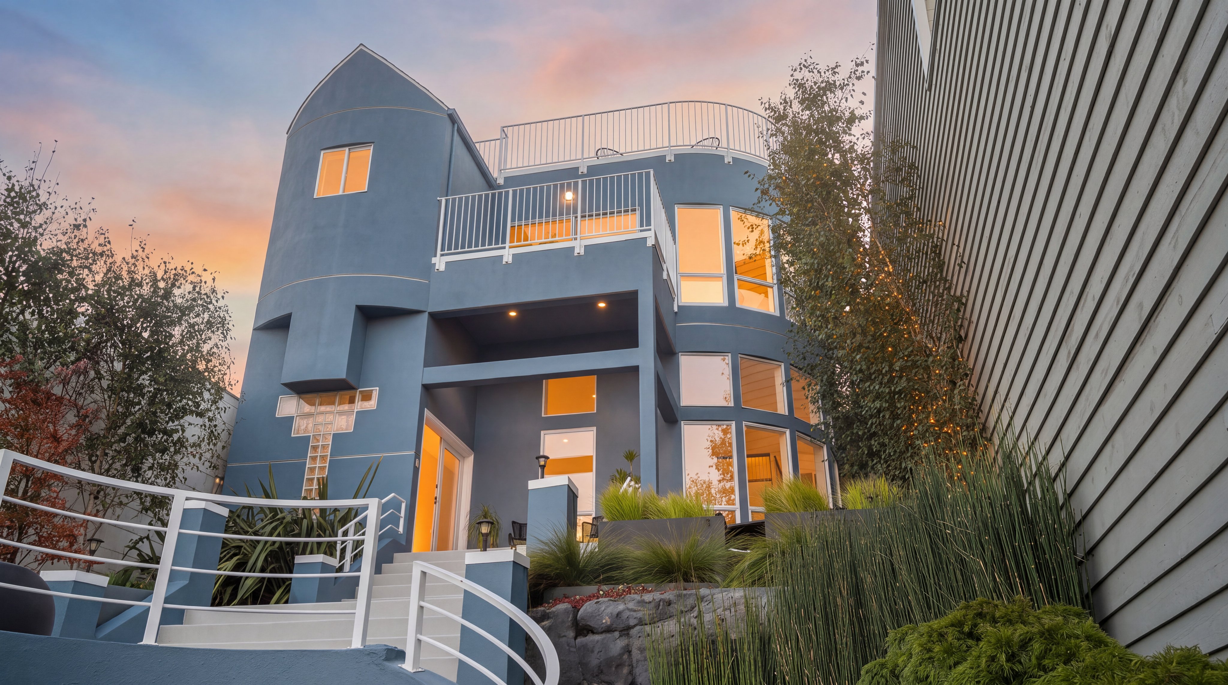 This striking contemporary home features a unique blue stucco facade with a prominent rounded tower element and a multi-level design. The exterior includes a rooftop deck with white railings, large windows that capture natural light, and a tiered landscape with stone retaining walls and lush greenery. A curved staircase with a white railing leads up to the main entrance, creating an inviting and architecturally distinct approach.