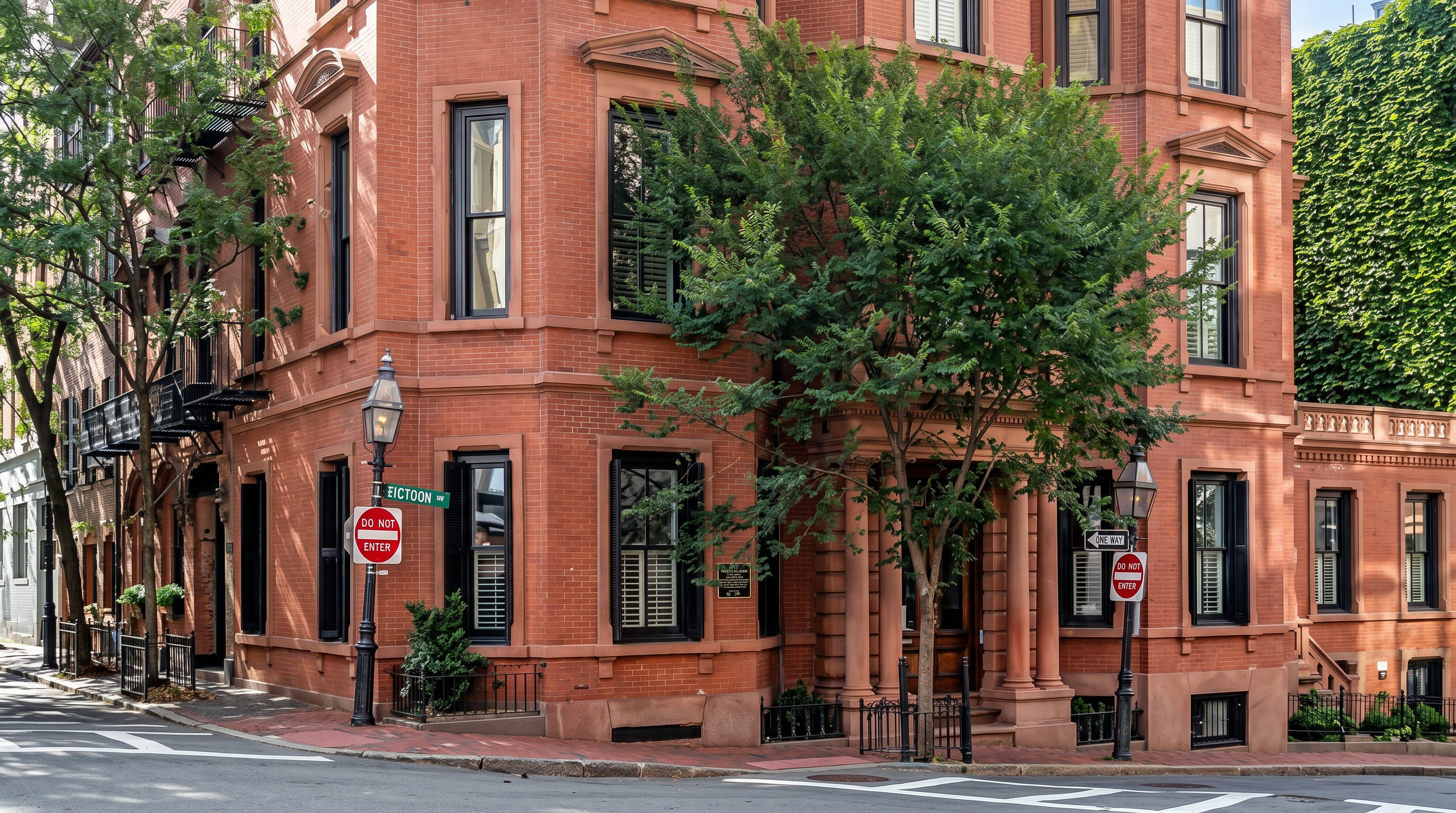 This striking multi-story brick building showcases classic Second Empire architectural style, characterized by its prominent mansard roof and ornate dormer windows. The structure occupies a prominent corner lot, featuring a rich red masonry facade, symmetrical window placement, and a stately entrance framed by columns. A mature tree partially obscures the lower levels, adding a touch of natural softness to the formal urban aesthetic.