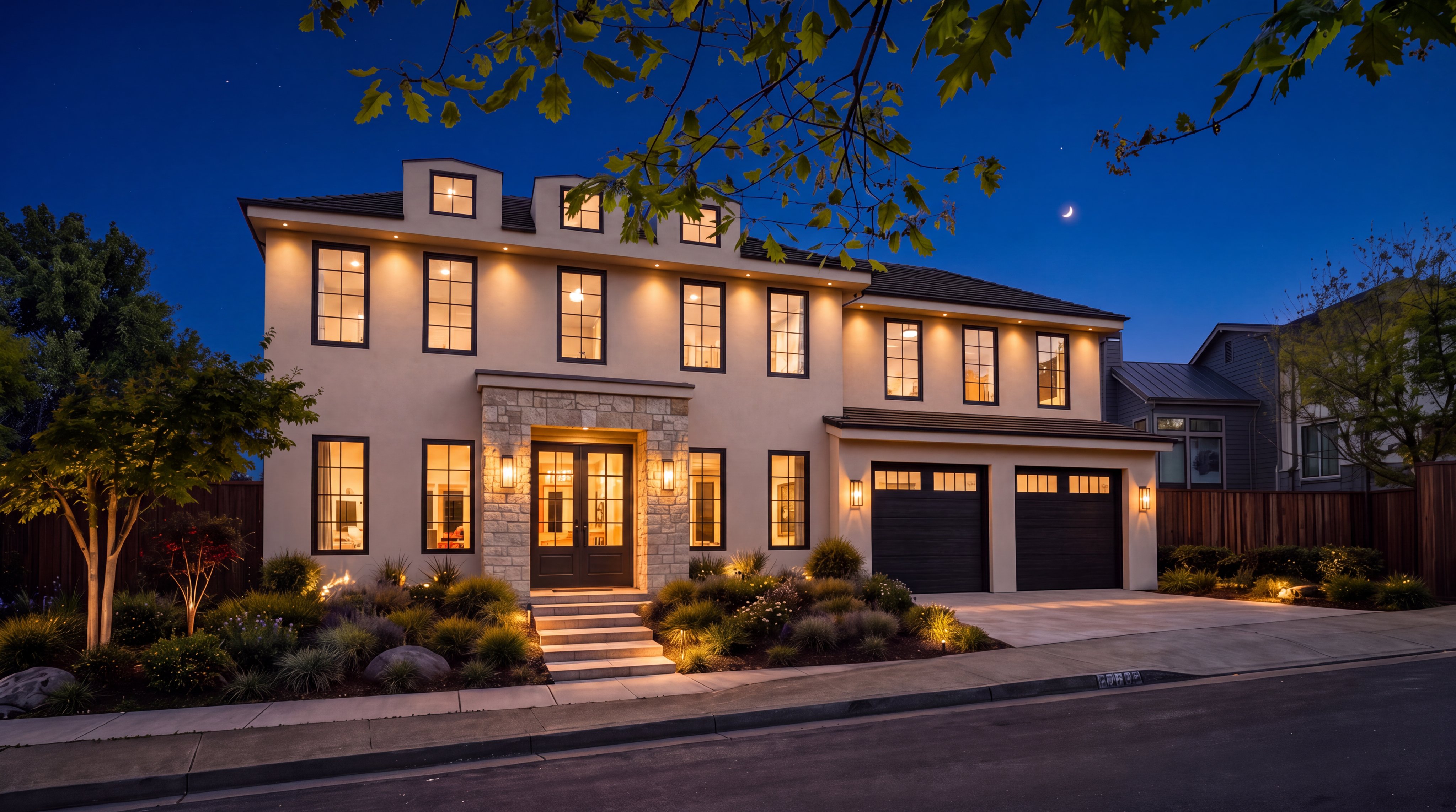 This elegant two-story residence features a sophisticated blend of light-colored stucco and natural stone accents, creating a timeless architectural aesthetic. The home boasts a symmetrically designed facade with black-trimmed windows, a grand double-door entrance, and a dual-car garage, all illuminated by warm, inviting outdoor lighting. Beautifully landscaped shrubbery and low-profile plantings enhance the curb appeal, making this an ideal showcase for a premium residential listing.