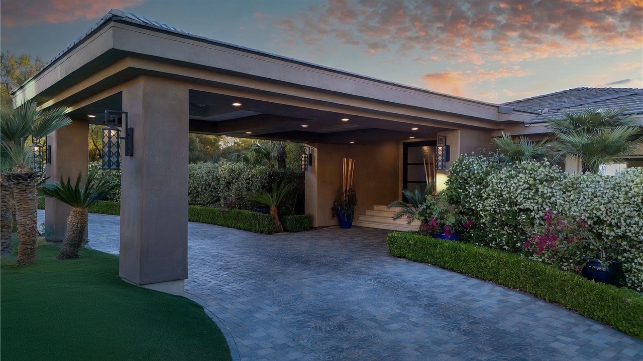 This elegant front entryway features a grand porte-cochère that provides a sheltered arrival area with recessed lighting. The driveway is paved with textured stone pavers, leading toward a modern glass-paneled front door framed by lush, manicured landscaping and flowering shrubs. The scene is captured at dusk, with the warm glow of exterior lanterns highlighting the sophisticated architectural design.