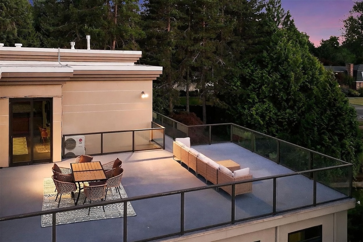This elevated rooftop deck offers a spacious outdoor living area featuring a dining set on a patterned rug and a long, modular sectional sofa. The space is enclosed by modern glass railings, providing an unobstructed view of the surrounding mature trees and evening sky. The perspective is captured from an elevated angle, highlighting the deck's layout and its seamless connection to the home's interior through sliding glass doors.