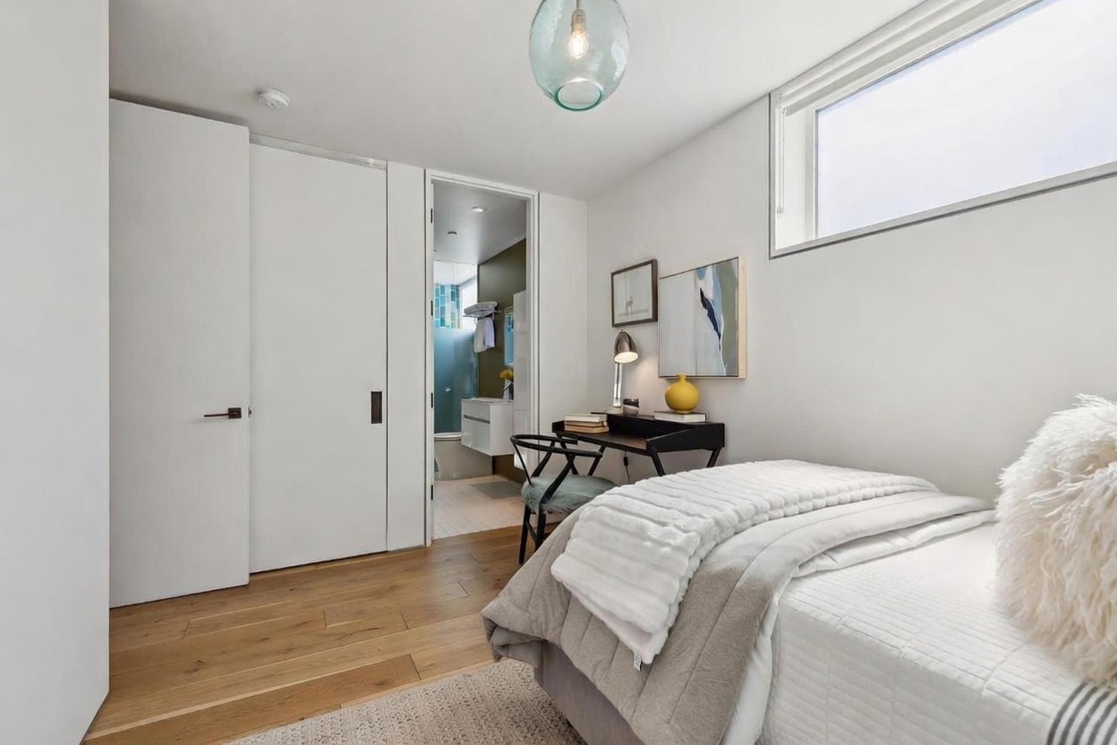This bright and modern bedroom features light wood flooring, a neutral color palette, and a cozy bed dressed in layered linens. A small desk area is tucked against the wall, complemented by a minimalist chair and wall art, while a doorway leads directly into an adjacent bathroom. The room is illuminated by a unique glass pendant light and natural light streaming through a high, horizontal window.