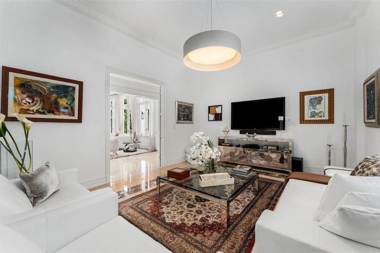 This elegant living room features a bright, neutral color palette with white sofas arranged around a central glass-topped coffee table and a detailed, traditional-style area rug. A large flat-screen television is mounted above a mirrored media console, complemented by various framed artworks on the walls. The space is illuminated by a large, modern drum pendant light and opens into an adjacent sunlit room through a wide doorway, creating an airy and sophisticated atmosphere.