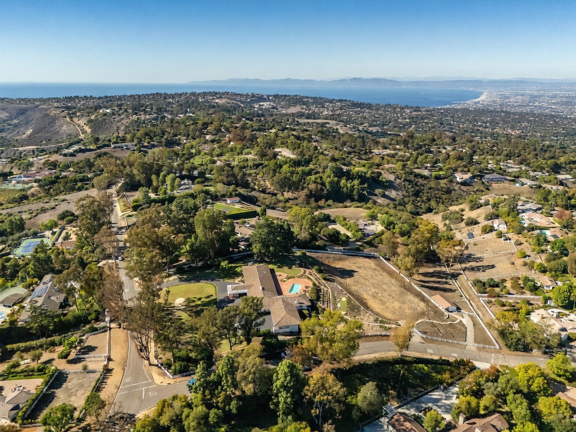 This high-angle aerial perspective captures a sprawling residential property nestled within a lush, hilly landscape overlooking the distant coastline. The estate features a large, multi-wing home with a swimming pool, surrounded by expansive grounds, mature trees, and winding roads. The wide-reaching view emphasizes the property's secluded setting and its proximity to the ocean, offering a sense of privacy and scenic beauty.