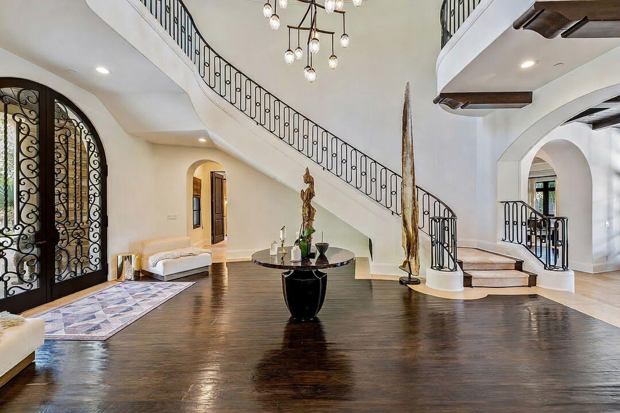 This grand foyer features a sweeping staircase with intricate wrought-iron railings and dark wood flooring that reflects the light from a modern, multi-tiered chandelier. A central circular table serves as a focal point, topped with a decorative sculpture, while an arched doorway and a large, ornate iron-framed glass door add architectural character. The space is bright and airy, conveying a sense of luxury and sophisticated design.