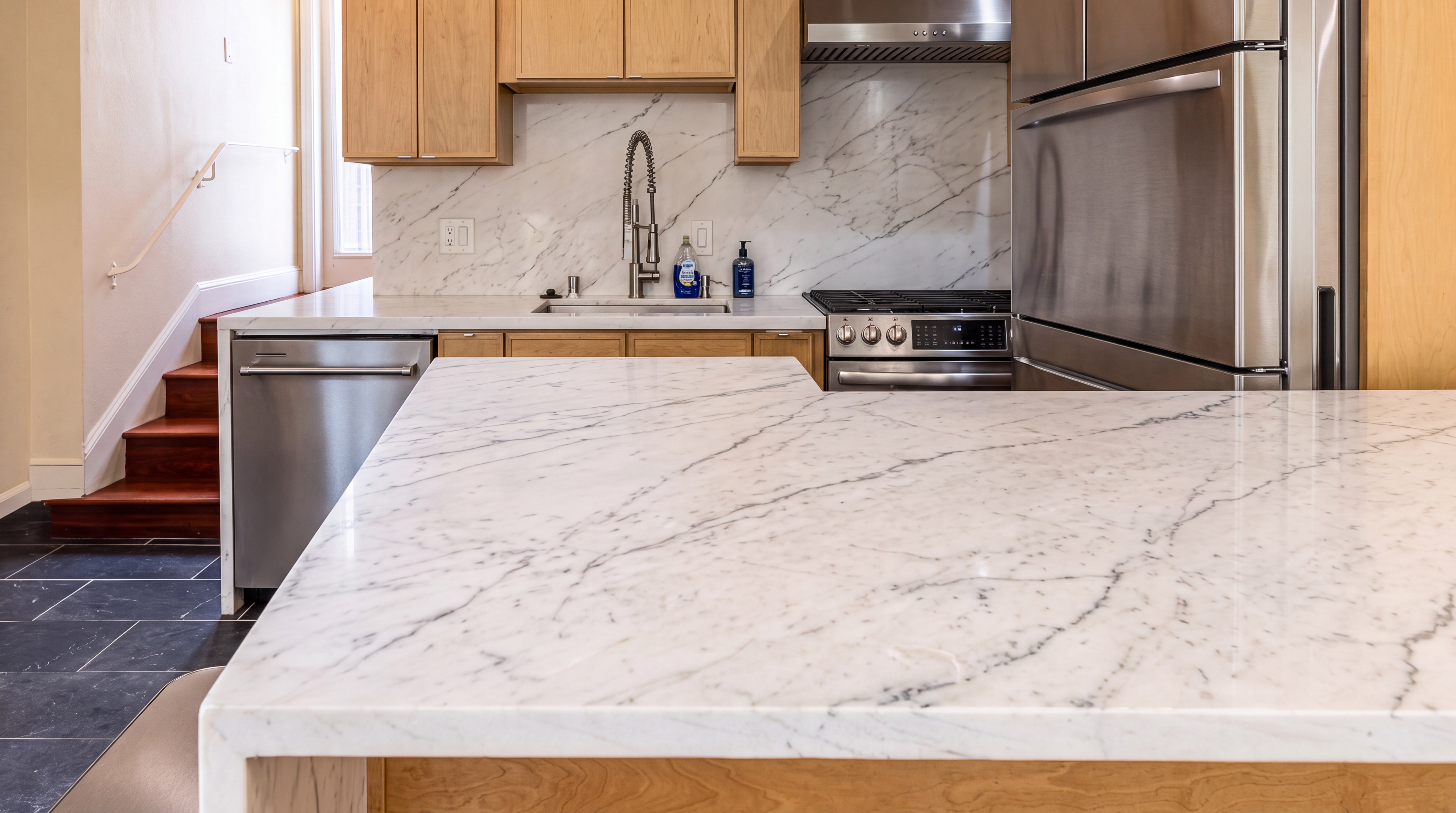 This kitchen features a clean, contemporary design with light wood cabinets, high-end stainless steel appliances, and elegant marble countertops. A large waterfall-edge kitchen island provides a spacious prep area, while natural stone surfaces extend to the backsplash for a cohesive look. The area also conveniently connects to a staircase, blending modern luxury with a functional layout.