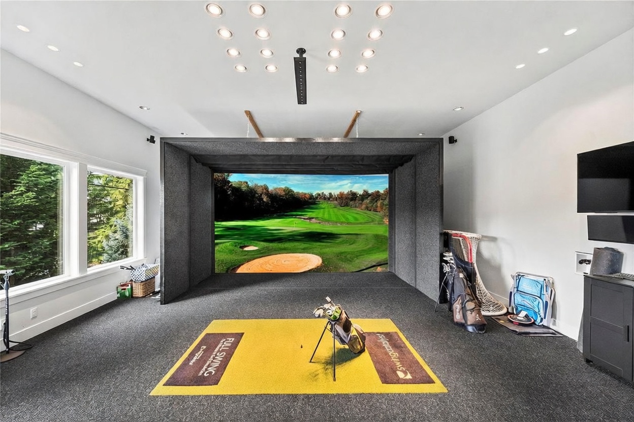 This dedicated home golf simulator room features a large, professional-grade impact screen housed within a dark, sound-dampening enclosure. The space is equipped with a yellow turf hitting mat, a golf bag, and integrated ceiling lighting, creating a focused environment for indoor practice. Large windows on the left side provide natural light and a view of the outdoors, while the neutral color palette and carpeted flooring maintain a clean, functional aesthetic.