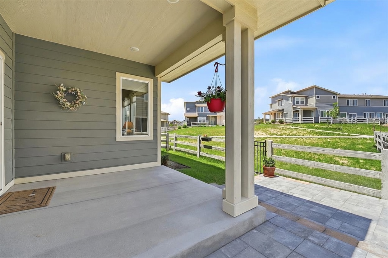 This covered patio features a smooth concrete floor and a clean, modern aesthetic with gray horizontal siding. A hanging flower basket adds a touch of color, while the open side offers a view of a grassy backyard, a white split-rail fence, and neighboring homes under a bright blue sky. The perspective is from the covered porch looking out toward the landscaped yard and surrounding residential area.