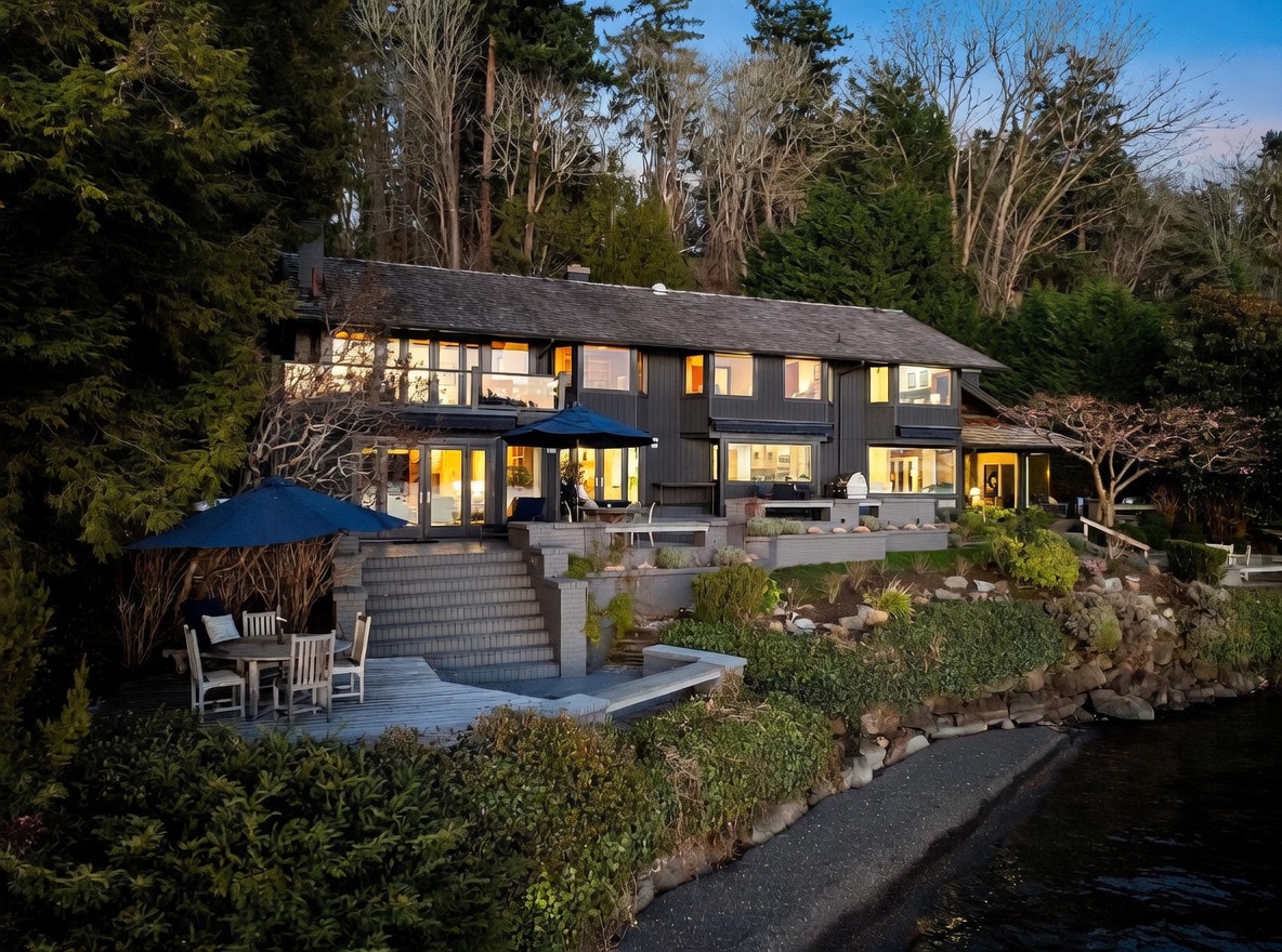 This stunning waterfront property features a multi-level dark-toned exterior with expansive windows and a spacious tiered patio area. The home is nestled against a lush, wooded hillside, offering a seamless transition between indoor living spaces and the natural shoreline. The scene is captured at dusk, with warm interior lighting creating an inviting and sophisticated atmosphere.