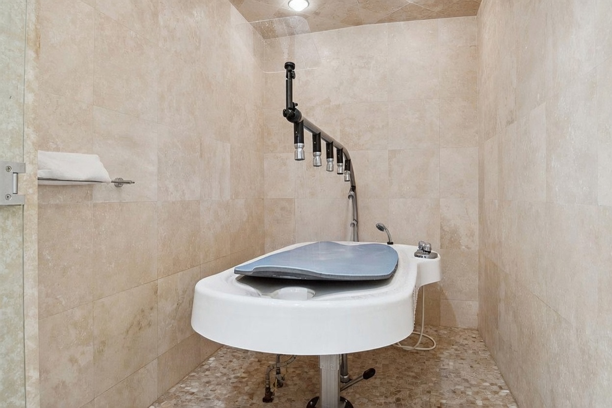 This specialized pet grooming station is located within a tiled shower enclosure, featuring a raised white basin with a flexible handheld sprayer and a multi-nozzle shower head system. The walls are finished in large, neutral-toned stone tiles, and the floor consists of small, textured pebble-style tiles. The space is designed for convenience and functionality, providing a dedicated area for pet care.