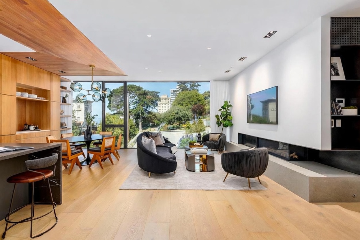 This open-concept living and dining area features light wood flooring and a warm wood-paneled ceiling accent that defines the dining space. The living area is anchored by a curved sofa, a pair of dark velvet armchairs, and a long, low-profile fireplace set into a concrete-style hearth. Large windows provide an expansive view of the lush greenery outside, while a modern multi-globe chandelier hangs above the dining table.
