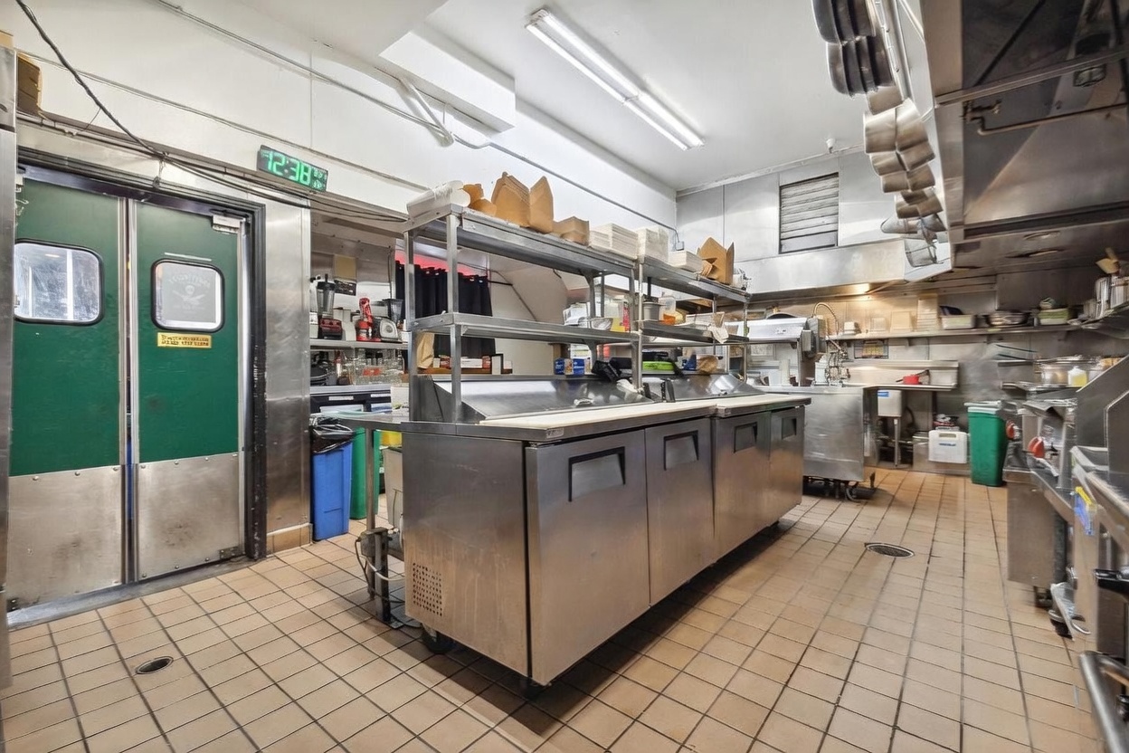 This commercial kitchen space features industrial-grade stainless steel prep tables, shelving units, and professional-grade appliances. The room is finished with tiled flooring, a drop ceiling with fluorescent lighting, and a green double-door entrance. The perspective is a wide-angle shot looking across the central prep station, highlighting the functional and utilitarian layout of a professional food service environment.