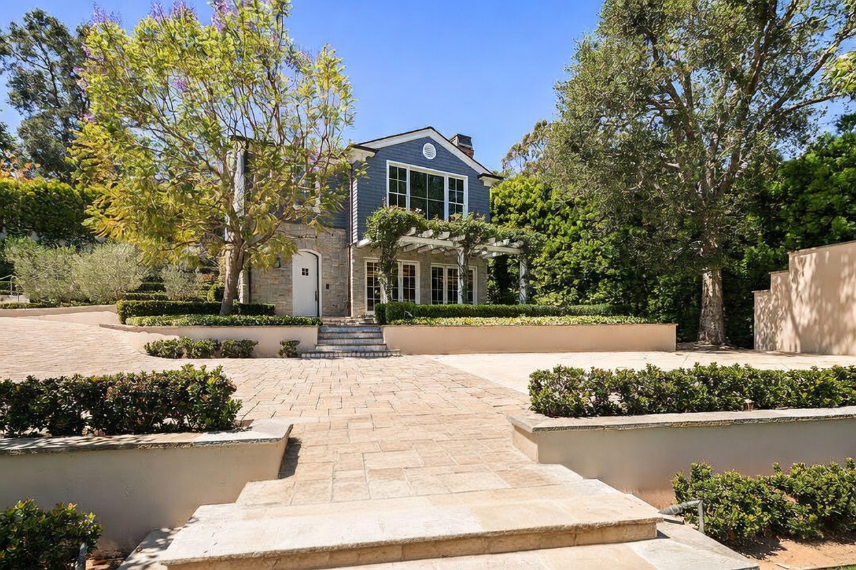 This elegant two-story residence features a blend of stone and blue-grey shingle siding, set against a backdrop of lush, mature trees. A wide, paved driveway leads to a welcoming front entrance, complemented by a tiered stone staircase adorned with decorative patterned tiles. The property includes a pergola-covered patio area and manicured landscaping, creating a sophisticated and inviting curb appeal.