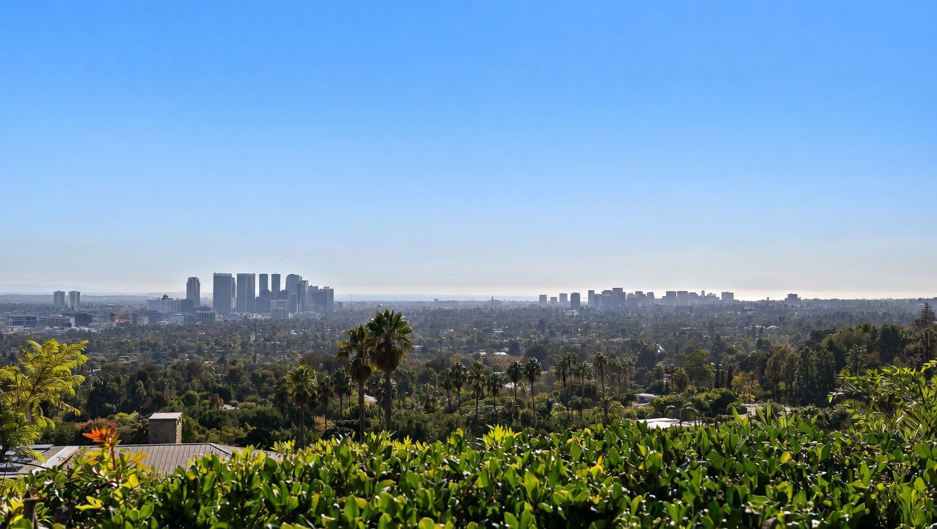 This image captures a breathtaking, elevated panoramic view of a sprawling cityscape from what appears to be a private balcony or terrace. The foreground features lush green foliage and palm trees, leading the eye toward a dense urban skyline set against a clear blue sky and a distant horizon line. The perspective offers a sense of luxury and tranquility, highlighting the property's prime location and expansive vistas.