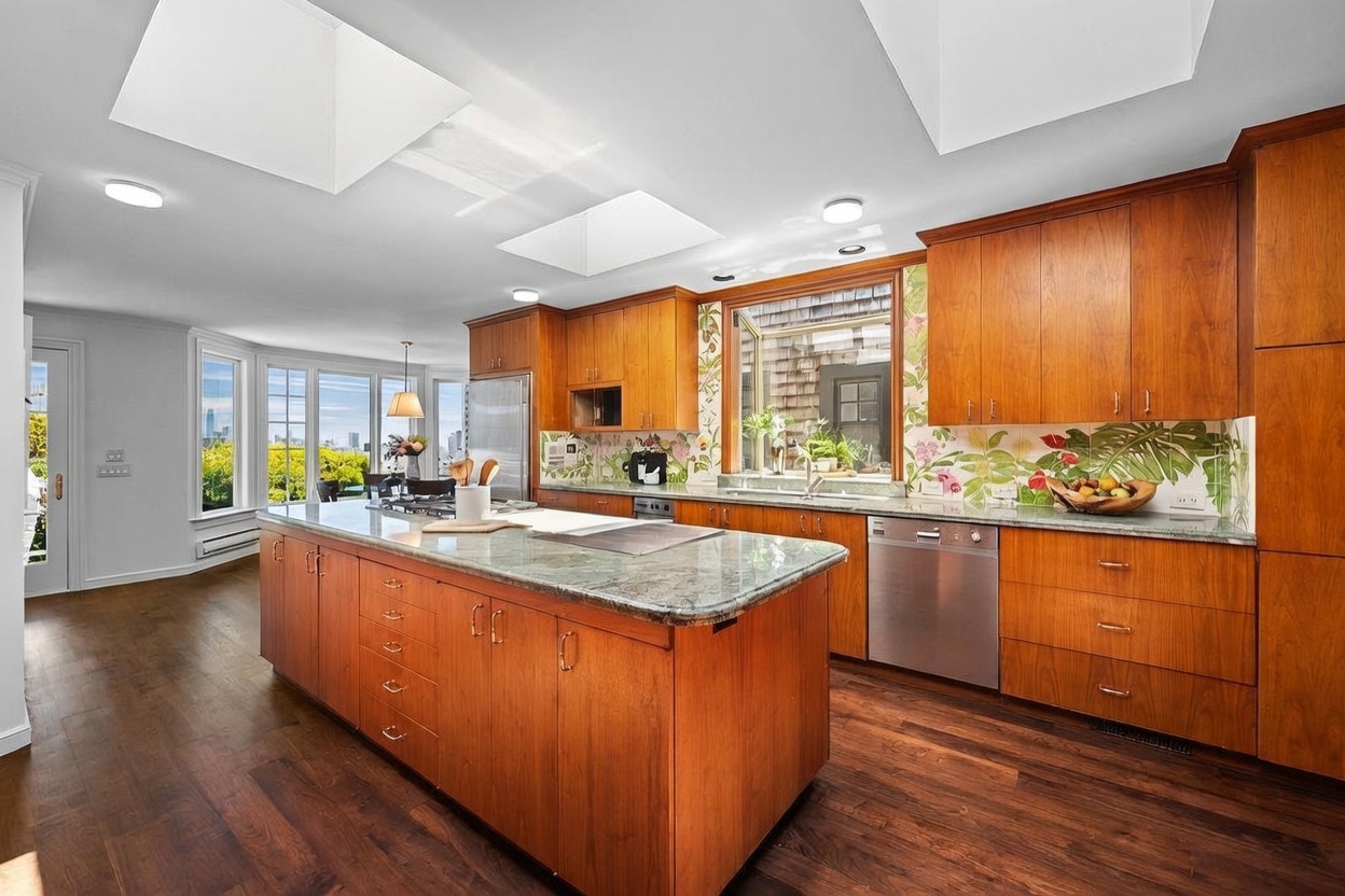 This spacious kitchen features warm-toned wood cabinetry, a large central island with a stone countertop, and a distinctive floral-patterned backsplash. The room is brightened by multiple skylights and large windows that provide a view of the outdoors. The perspective is a wide-angle shot from the kitchen floor, showcasing the open layout and the seamless transition between the cooking area and the adjacent dining space.