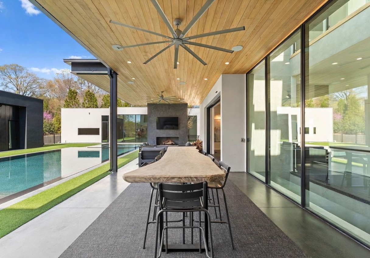 This modern covered patio features a natural-edge wood dining table set against a backdrop of a sleek swimming pool and minimalist architecture. The space is defined by a warm wood-paneled ceiling with a large fan and a tall stone-clad chimney breast housing an outdoor fireplace. Large glass panels provide a seamless transition between the indoor living area and the expansive outdoor terrace.