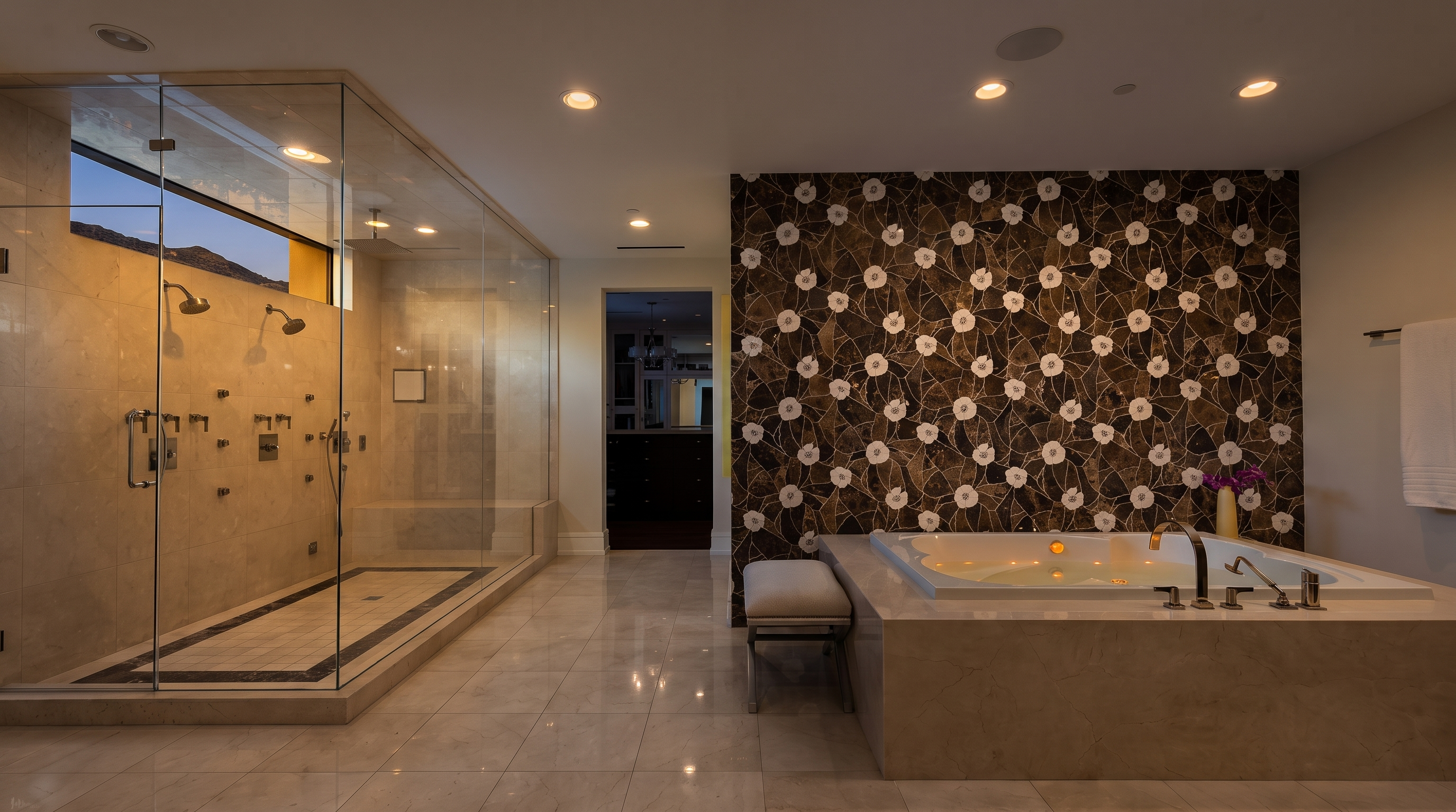 This luxurious primary bathroom features a large glass-enclosed walk-in shower with multiple showerheads and a separate soaking tub set against a striking, dark-toned mosaic tile wall with a floral pattern. The space is finished with light-colored floor tiles and recessed lighting, creating a bright and sophisticated atmosphere. A small bench sits beside the tub, and a doorway in the background leads to an adjacent room, providing a sense of depth and flow.
