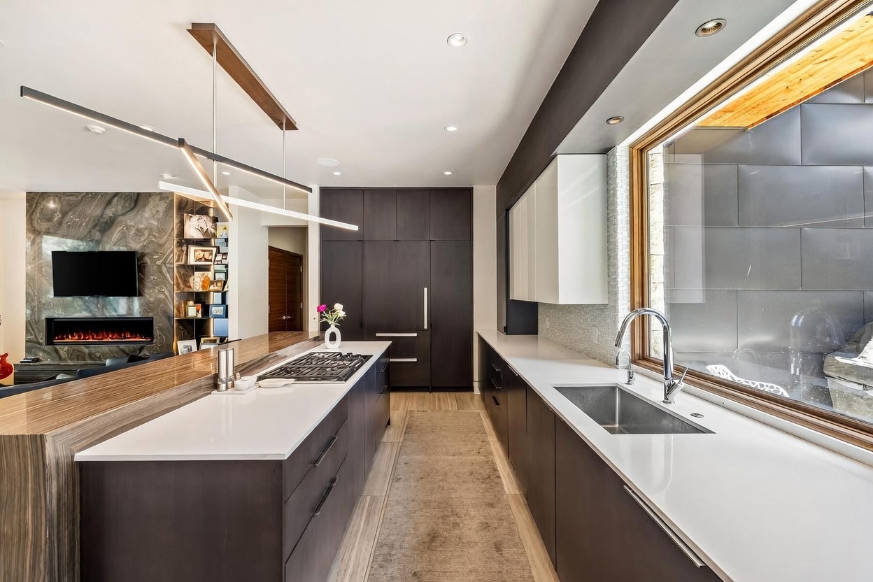 This modern kitchen features a long central island with a gas cooktop and a sleek, dark-toned cabinetry suite. A large window above the sink provides ample natural light, while a contemporary linear pendant light hangs above the island. The space flows seamlessly into an adjacent living area, which is anchored by a fireplace with a tall, textured stone surround.