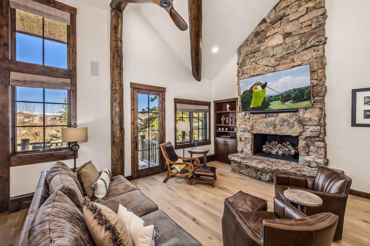 This rustic living room features a vaulted ceiling with exposed timber beams and a prominent stone-clad chimney breast housing a television above the firebox. The space is furnished with a plush brown sofa, leather armchairs, and a wooden coffee table, all arranged on light wood flooring. Large windows provide ample natural light and views of the surrounding landscape, creating a warm and inviting mountain-lodge atmosphere.
