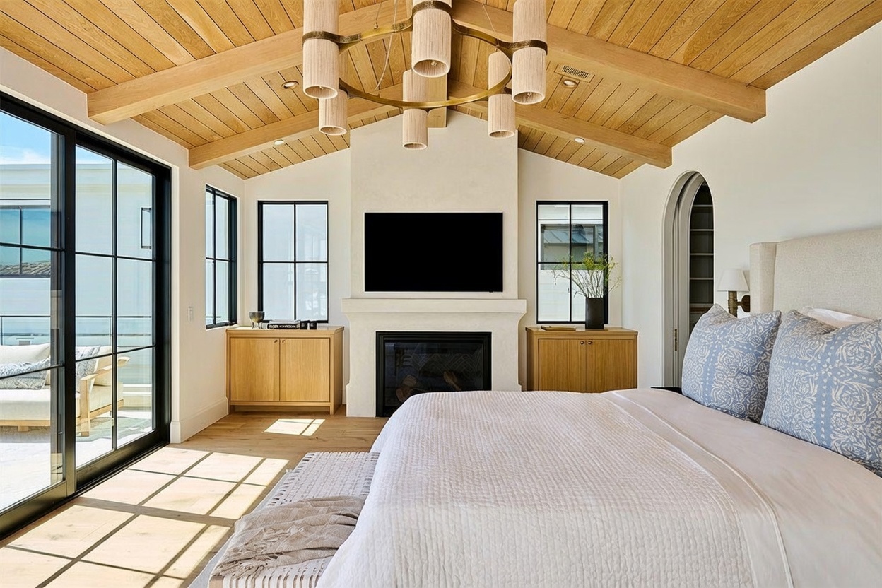 This bright and airy primary bedroom features a vaulted wood-paneled ceiling with a large, multi-tiered chandelier as a central focal point. A fireplace with a tall, light-colored surround sits between two wooden cabinets, flanked by windows that look out onto a balcony. The room is furnished with a large bed featuring patterned pillows and offers a seamless transition to the outdoor space through expansive glass sliding doors.