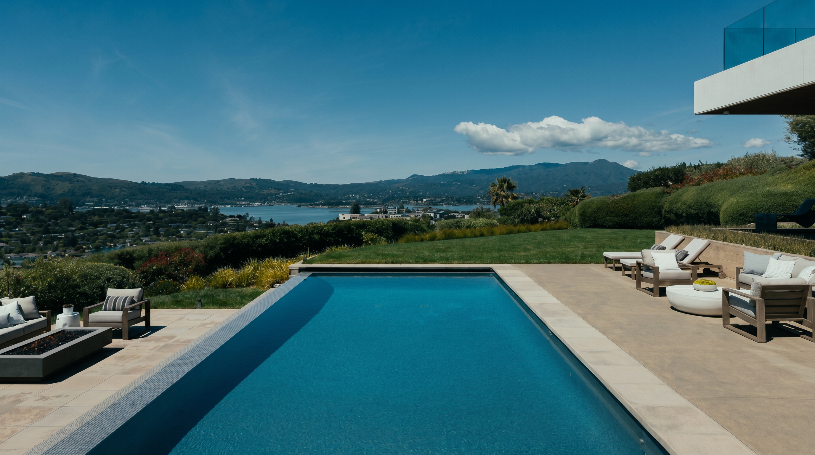 This stunning outdoor space features a sleek, rectangular infinity-edge pool that overlooks a sprawling bay and distant mountain range. The expansive stone patio is furnished with modern lounge chairs and a fire pit area, creating an ideal setting for relaxation and entertaining. The scene is framed by lush green landscaping and a clear blue sky, emphasizing the property's luxurious and serene atmosphere.
