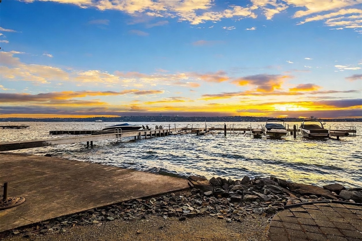 This scenic waterfront view captures a concrete patio area transitioning into a rocky shoreline overlooking a calm lake at sunset. Several private boat docks extend into the water, with multiple boats moored, creating a peaceful and recreational atmosphere. The vibrant orange and blue sky reflects across the water, emphasizing the property's prime location and serene outdoor living potential.