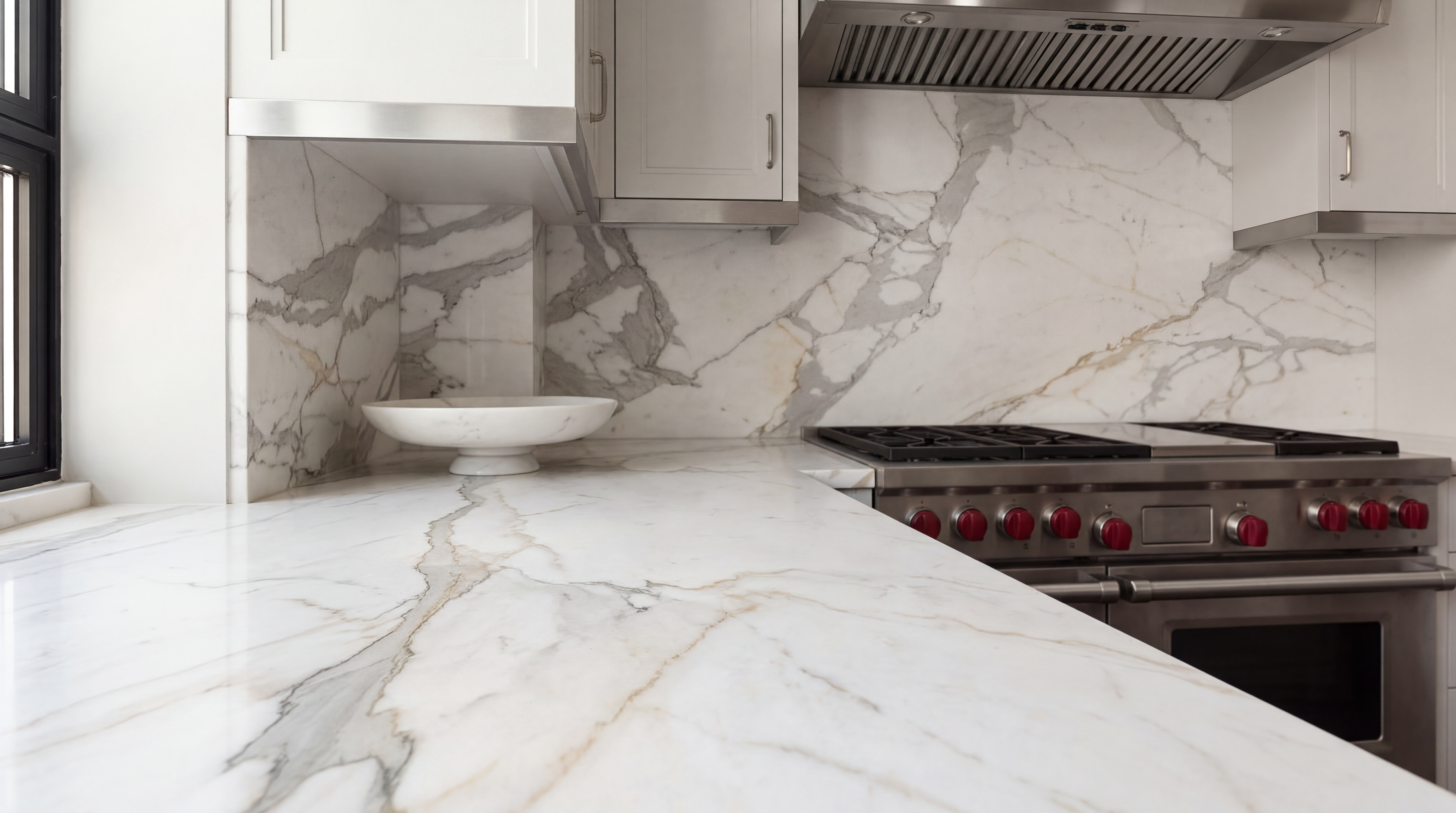 This sophisticated kitchen features stunning marble countertops and a matching full-height marble backsplash that adds a high-end, cohesive feel. The space is equipped with professional-grade stainless steel appliances, including a range with distinctive red knobs, and clean-lined cream cabinets. The overall aesthetic is elegant, timeless, and minimalist, emphasizing luxury finishes.