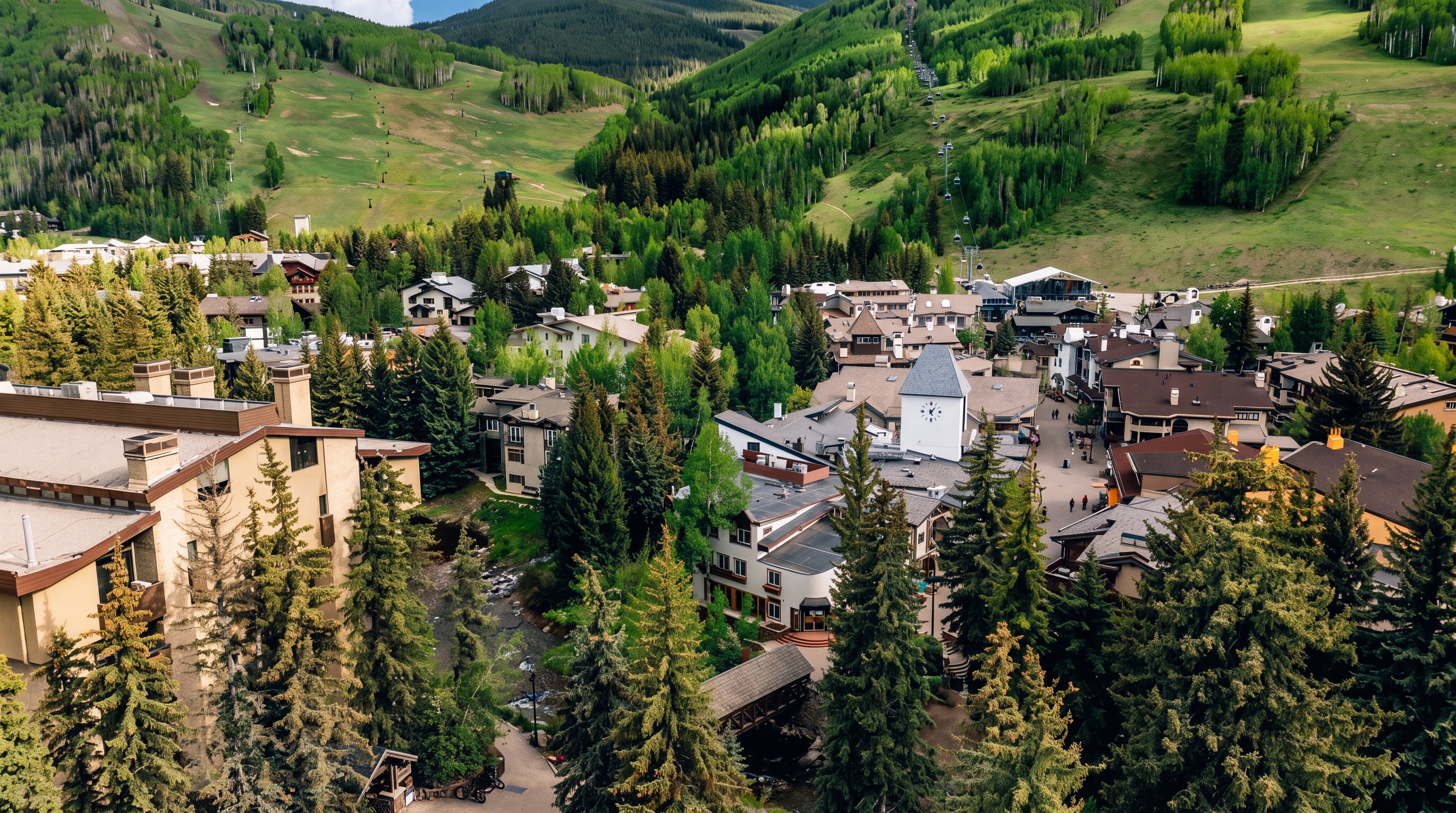 This high-angle aerial view captures a picturesque mountain resort village nestled at the base of lush, green ski slopes. The scene features a mix of multi-story residential buildings with wood and stone facades, surrounded by dense pine trees and a winding stream. The perspective provides a comprehensive look at the community layout, highlighting the proximity to the mountain terrain and the charming, alpine-inspired architecture.