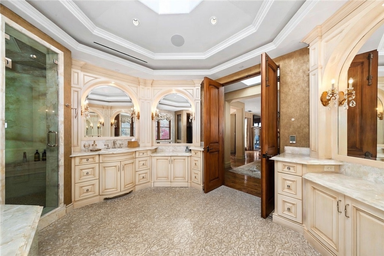 This spacious primary bathroom features a double vanity with light-toned cabinetry and stone countertops, complemented by arched mirrors and elegant wall sconces. To the left, a glass-enclosed shower stall is visible, while the room is finished with patterned carpeting and a high, multi-faceted ceiling. The open doorway leads into an adjacent living space, creating a sense of flow and luxury throughout the suite.
