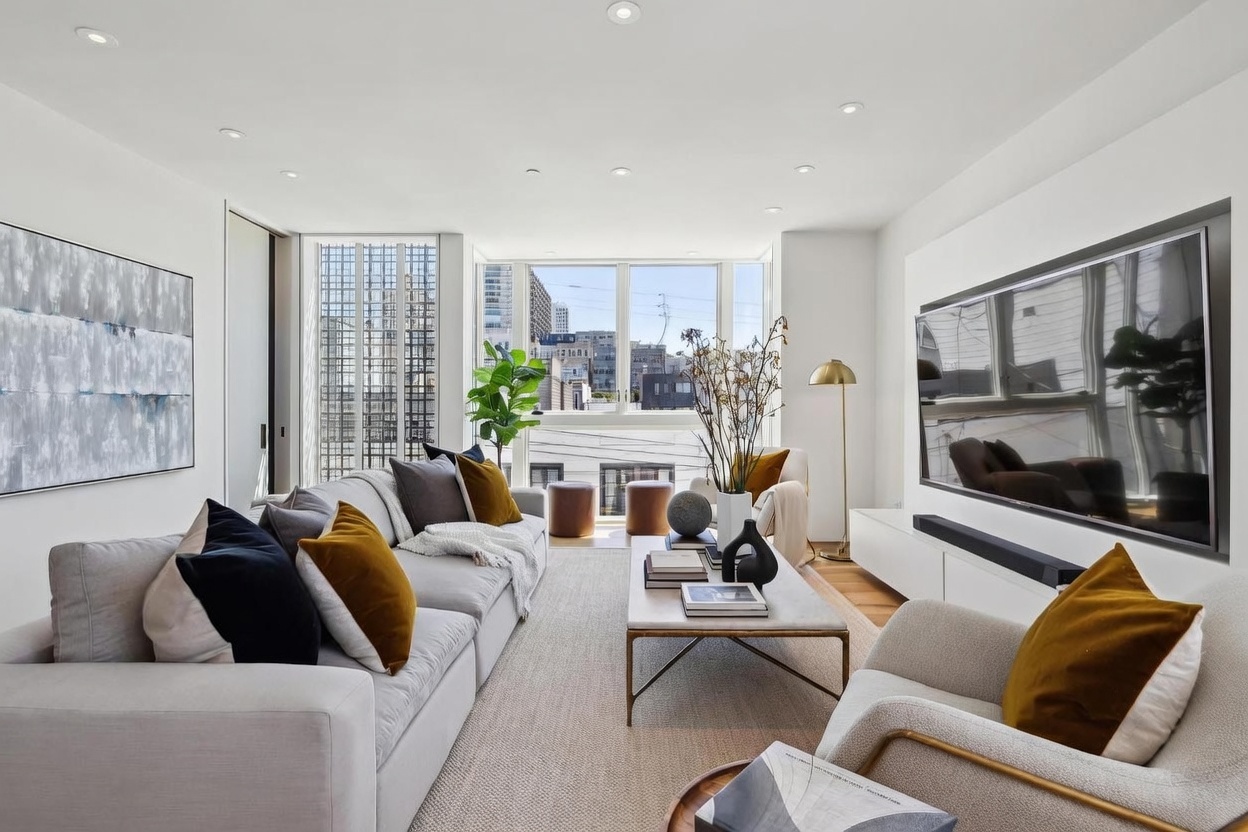 This bright and contemporary living room features a large, light-toned sectional sofa accented with dark and mustard-colored throw pillows. A central stone-topped coffee table sits on a textured area rug, while a large wall-mounted television serves as a focal point above a sleek, white media console. The space is illuminated by recessed lighting and natural light streaming through large windows that offer an urban view, creating an inviting and sophisticated atmosphere.