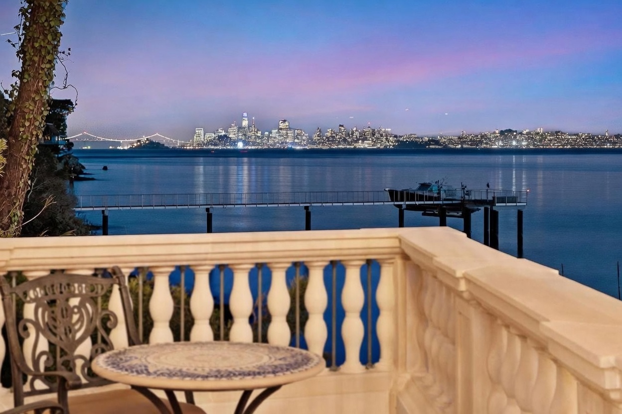 This elevated balcony offers a stunning, unobstructed panoramic view of the San Francisco skyline and the bay at dusk. The foreground features a classic stone balustrade and a small, round bistro table with a patterned top, creating an intimate setting for enjoying the scenery. A private pier extends into the calm water below, enhancing the serene and luxurious atmosphere of this waterfront property.