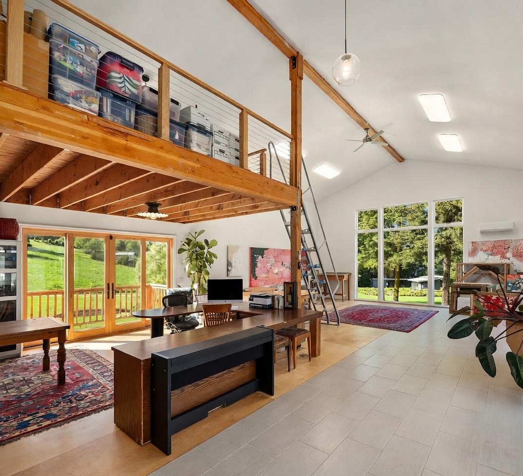This spacious, open-concept home office features a high, vaulted ceiling with exposed wooden beams and a lofted storage area accessible by a metal ladder. The room is filled with natural light from large windows and glass doors that provide a view of the surrounding greenery, while a desk setup occupies the center of the room. The space combines a rustic aesthetic with modern functionality, highlighted by light-toned flooring and a large, vibrant piece of wall art.