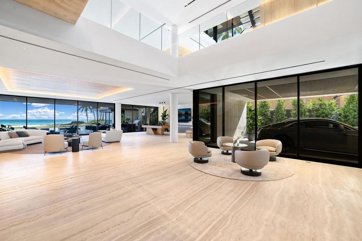 This expansive, open-concept living area features polished light-toned stone flooring and a dramatic double-height ceiling that enhances the sense of scale. The space is anchored by a seating arrangement on the left and a secondary conversation area on the right, all framed by expansive glass walls that provide views of the outdoors and a car displayed within the interior gallery space. The minimalist aesthetic is defined by clean lines, neutral furnishings, and a bright, airy atmosphere.