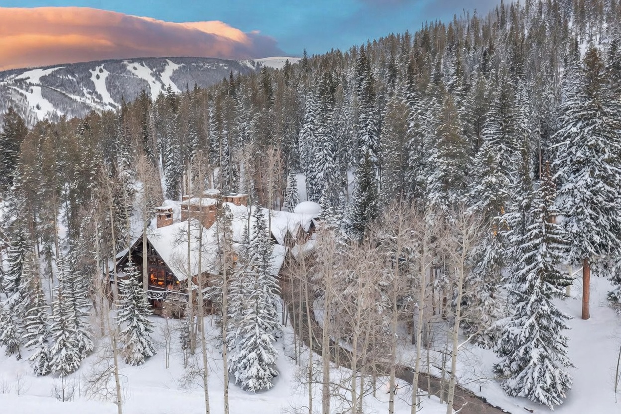 This aerial perspective captures a rustic, multi-story mountain estate nestled within a dense, snow-covered pine forest. The home features a complex roofline with multiple gables and stone-clad exterior walls, blending seamlessly into the winter landscape. In the background, a ski slope is visible on the distant mountain ridge, emphasizing the property's prime location in a high-altitude resort area.