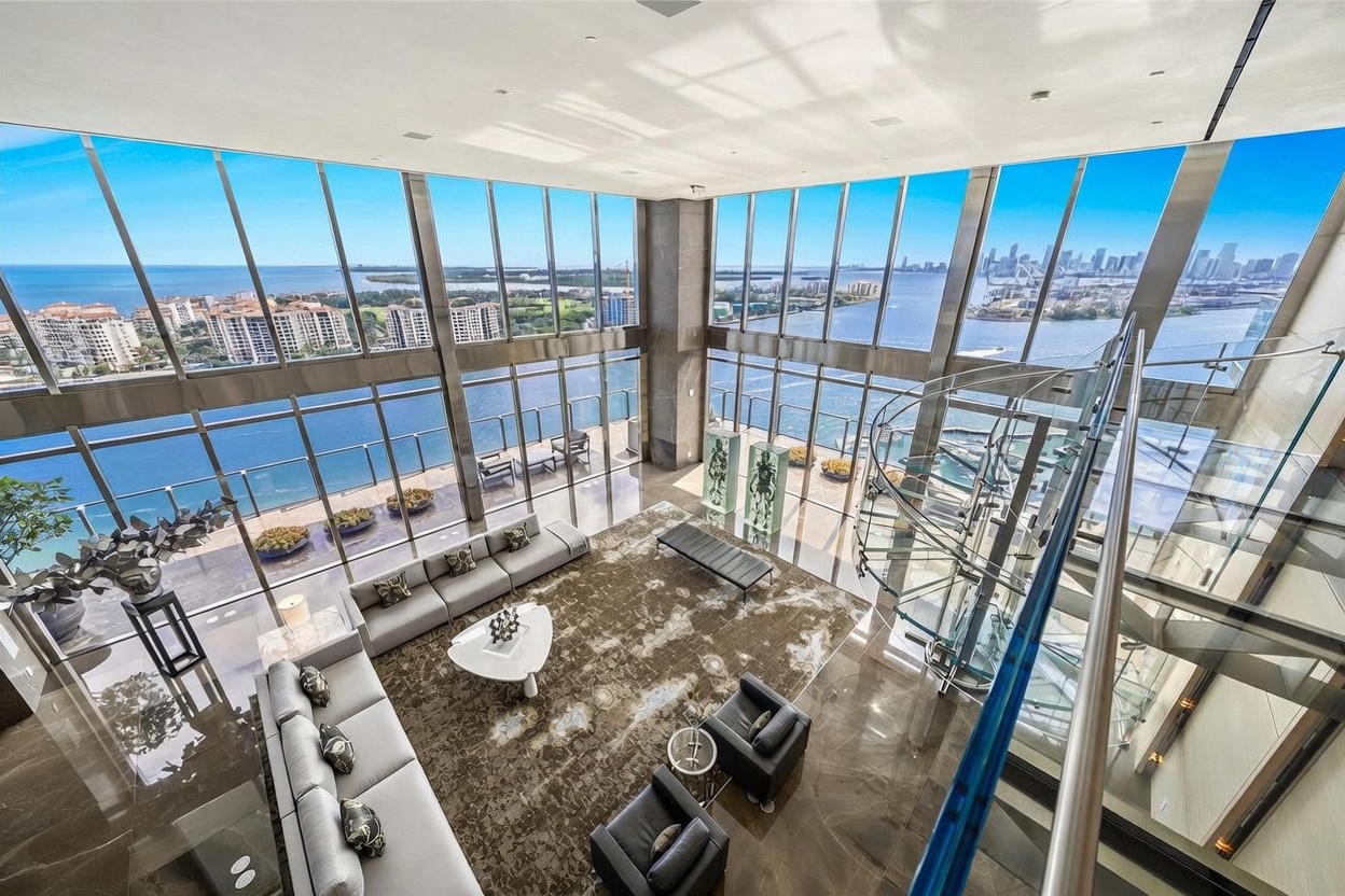 This expansive, double-height living room features dramatic floor-to-ceiling windows that offer panoramic views of the bay and city skyline. The space is anchored by a large, textured area rug and a neutral-toned sectional sofa, complemented by a sculptural white coffee table and dark leather armchairs. A striking glass-enclosed spiral staircase adds a modern architectural element, while the polished stone flooring reflects the abundant natural light filling the room.