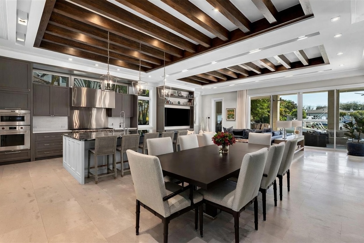This open-concept space features a large kitchen with a central island, dark cabinetry, and stainless steel appliances, seamlessly flowing into a formal dining area. The room is defined by a striking coffered ceiling with dark wood beams and recessed lighting, while large glass sliding doors provide a view of the outdoor area. The neutral color palette and clean lines create a sophisticated and airy atmosphere suitable for modern entertaining.