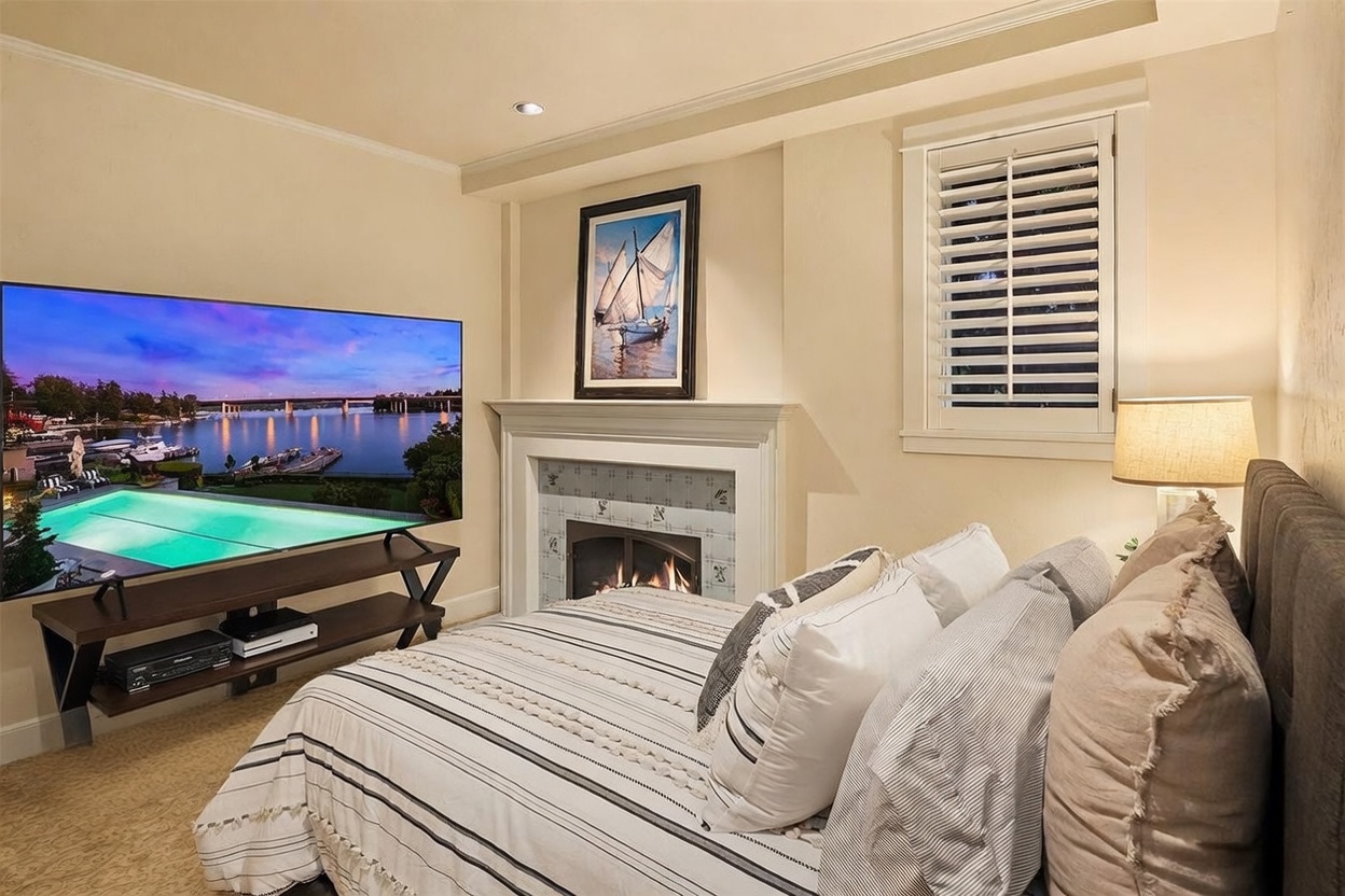This cozy bedroom features a neutral color palette and a comfortable bed dressed in striped bedding with decorative pillows. A large television sits on a dark wood media console, positioned across from a fireplace with a decorative mantel and a framed sailboat print above it. The room is illuminated by a bedside lamp and recessed lighting, creating a warm and inviting atmosphere.