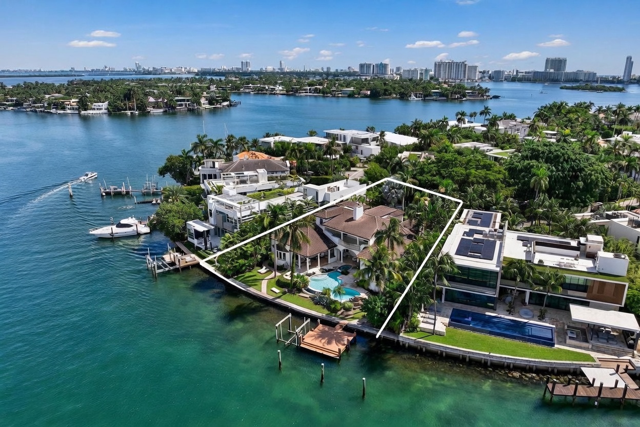 This high-angle aerial view captures a luxurious waterfront estate situated on a point, highlighted by a white outline. The property features a sprawling residence with a tiled roof, a private swimming pool, and a wooden dock extending into the turquoise bay. The surrounding landscape is lush with tropical greenery, and the background showcases a panoramic view of the city skyline across the water.