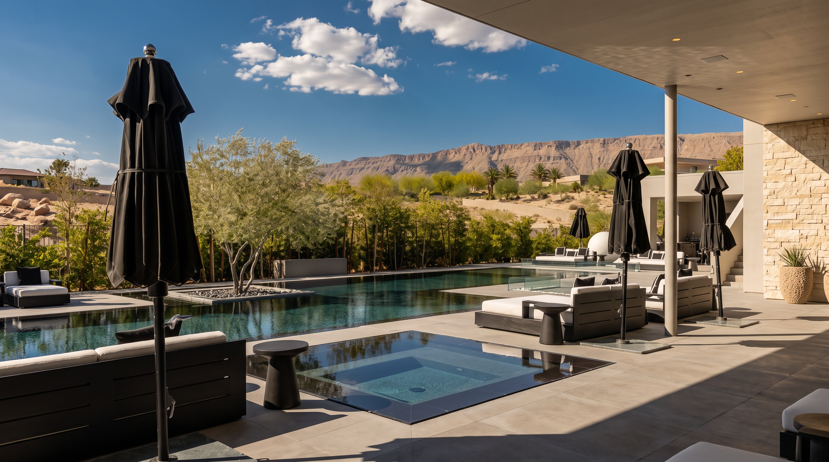 This luxurious outdoor living space features a sleek, modern infinity-edge pool and a square spa integrated into the patio deck. The area is furnished with contemporary lounge seating under black umbrellas, set against a backdrop of lush greenery and a dramatic desert mountain range. The perspective is from a covered patio, emphasizing the seamless transition between indoor and outdoor living.