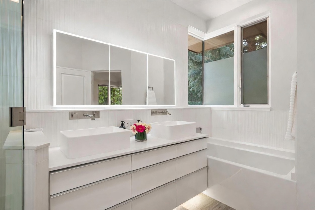 This modern primary bathroom features a sleek double vanity with white rectangular vessel sinks and wall-mounted faucets. A large, illuminated mirror spans the width of the vanity, reflecting the bright, minimalist space, while a deep soaking tub sits adjacent to a corner window. The room is finished with clean white surfaces and vertical textured wall tiles, creating a bright and contemporary atmosphere.