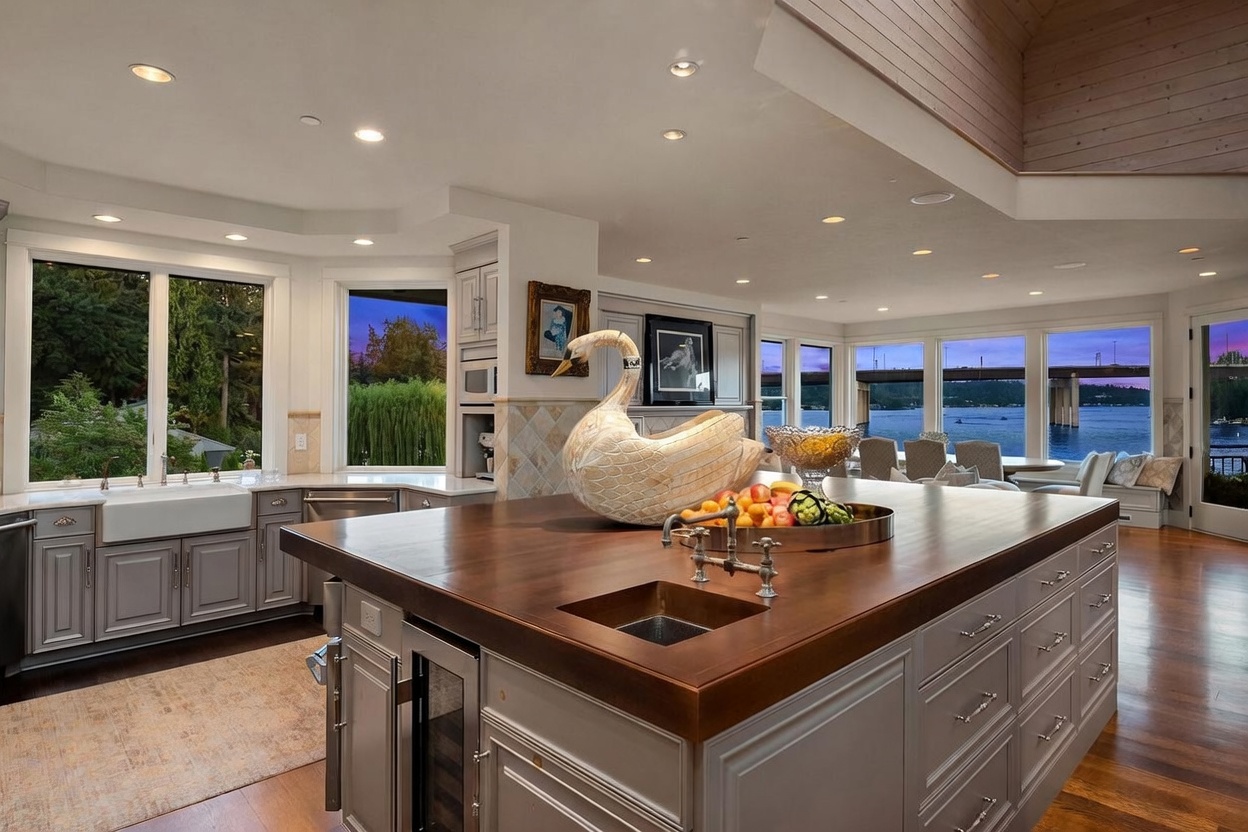 This spacious, high-end kitchen features a large central island with a dark wood countertop and a built-in prep sink, serving as the focal point of the room. The perimeter cabinetry is finished in a soft grey, complemented by a farmhouse-style sink and expansive windows that offer panoramic views of the water and bridge outside. The open-concept layout flows seamlessly into a dining area, creating an airy and sophisticated atmosphere perfect for entertaining.